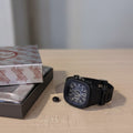 Black wristwatch on a wooden surface with a box in the background
